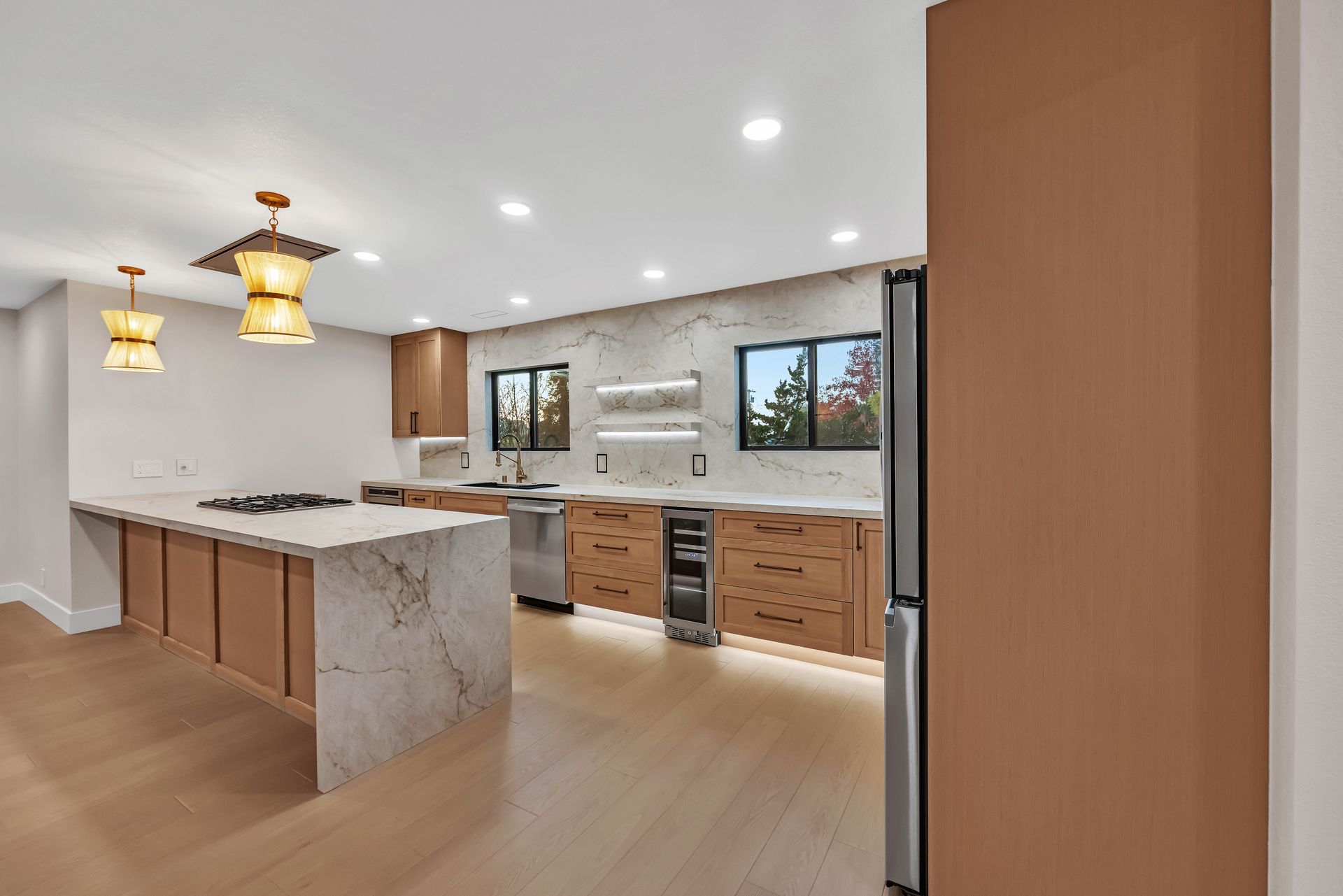 Modern kitchen with light wood cabinets, marble countertops, stainless steel appliances, and two pendant lights.