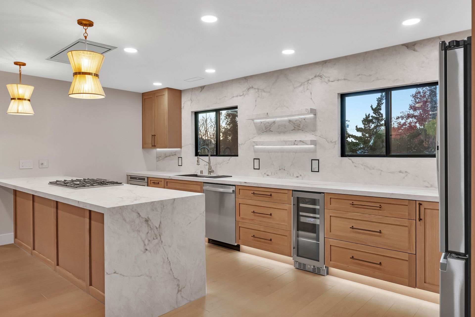 Modern kitchen with light wood cabinets, marble countertops, stainless steel appliances, and large windows.