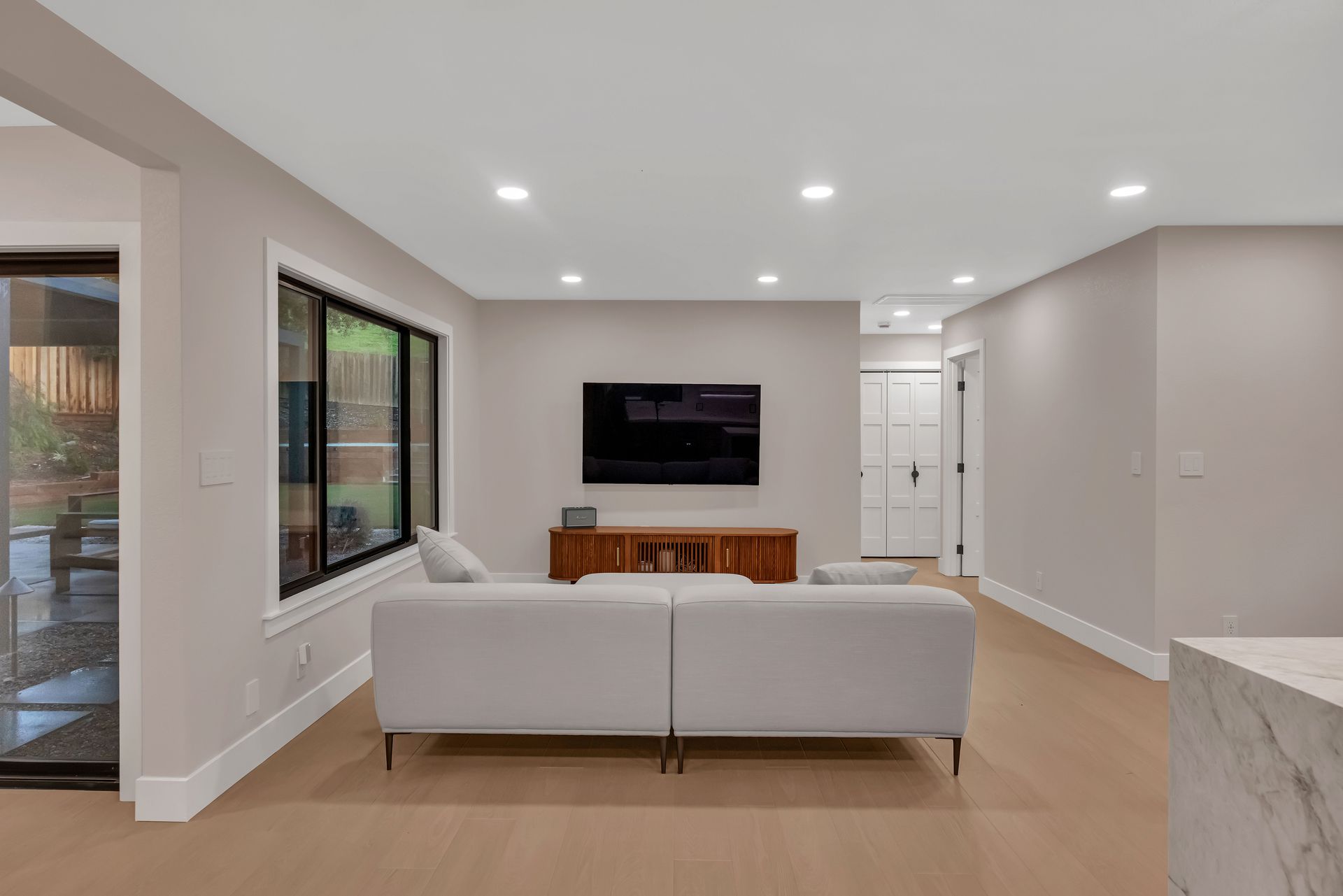 Living room with light-colored walls, sofa, TV on wooden console, and large window.