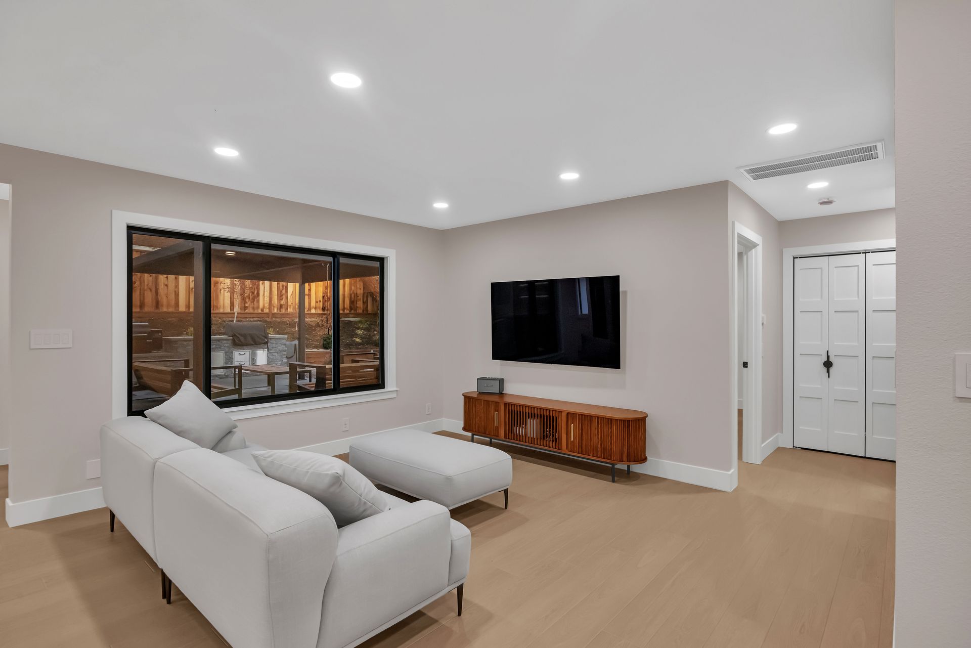 Modern living room with light walls, a couch, TV, and wooden media console. Large window.