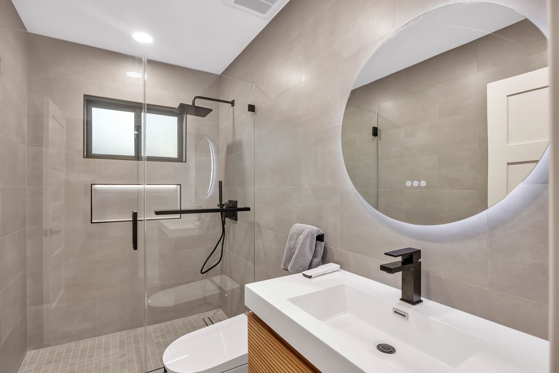 Modern bathroom with gray tiled walls, black fixtures, glass shower, and round mirror.