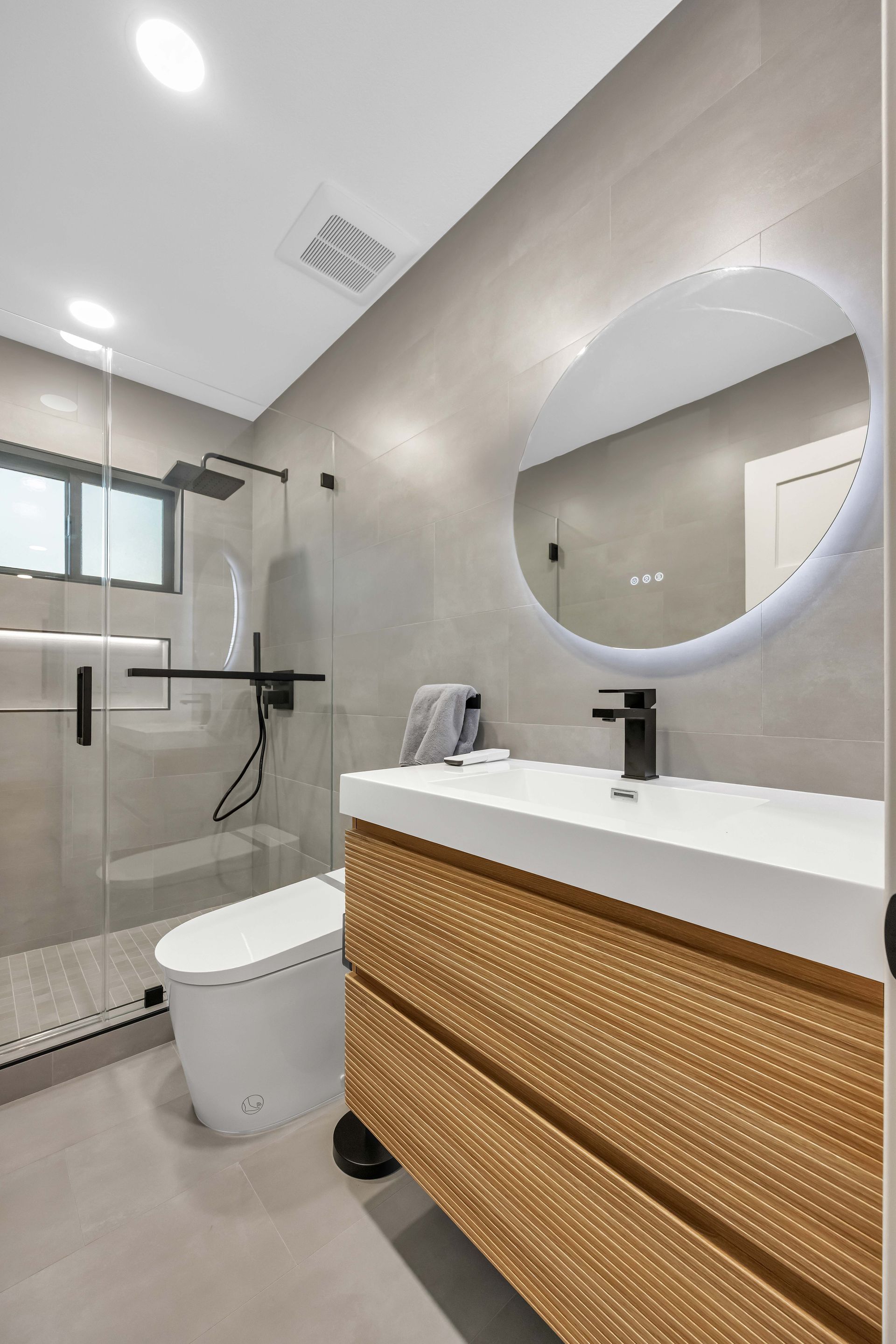 Modern bathroom with gray tiled walls, glass shower, wooden vanity, and oval mirror.