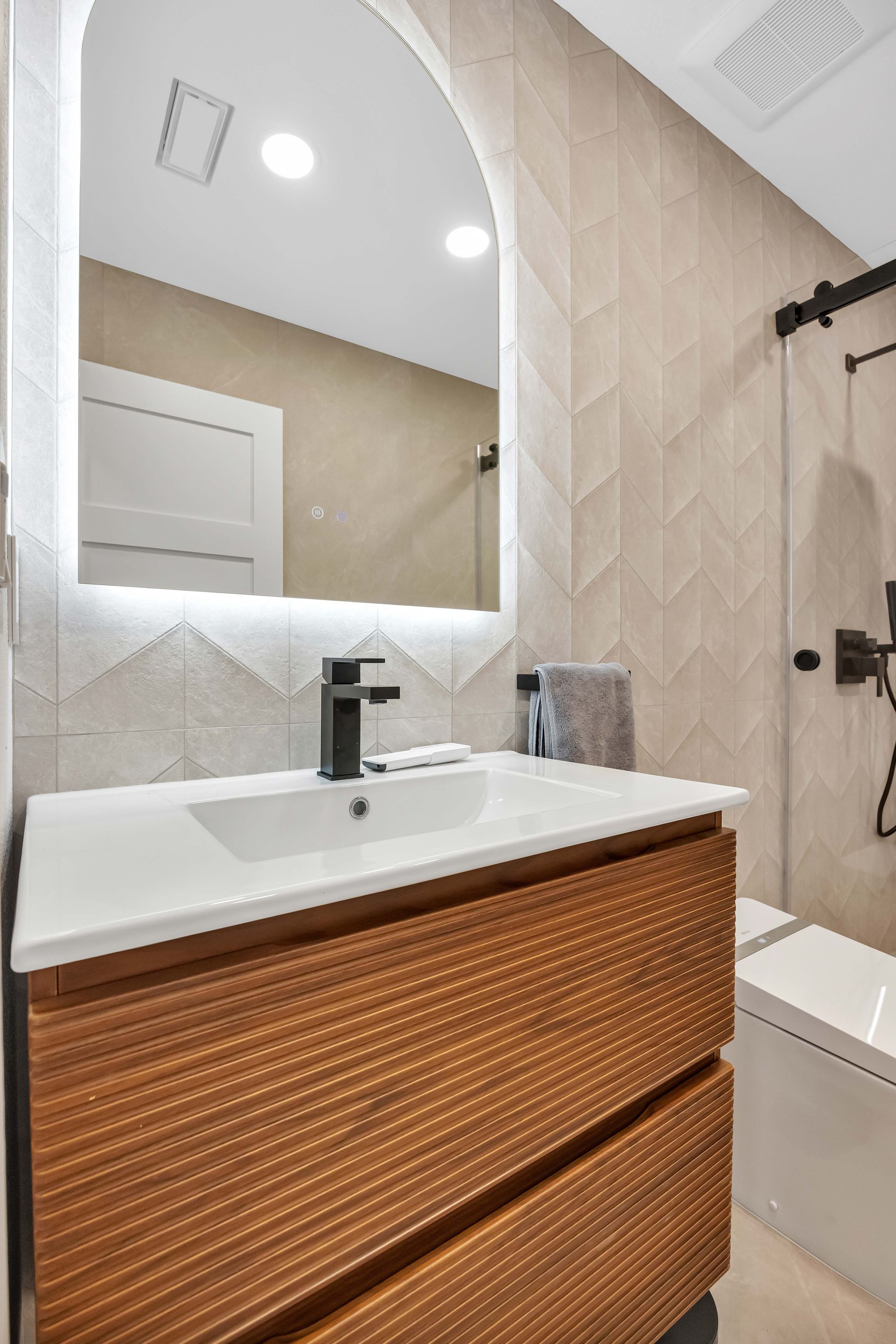 Modern bathroom with wood vanity, white countertop, and arched mirror with backlighting.