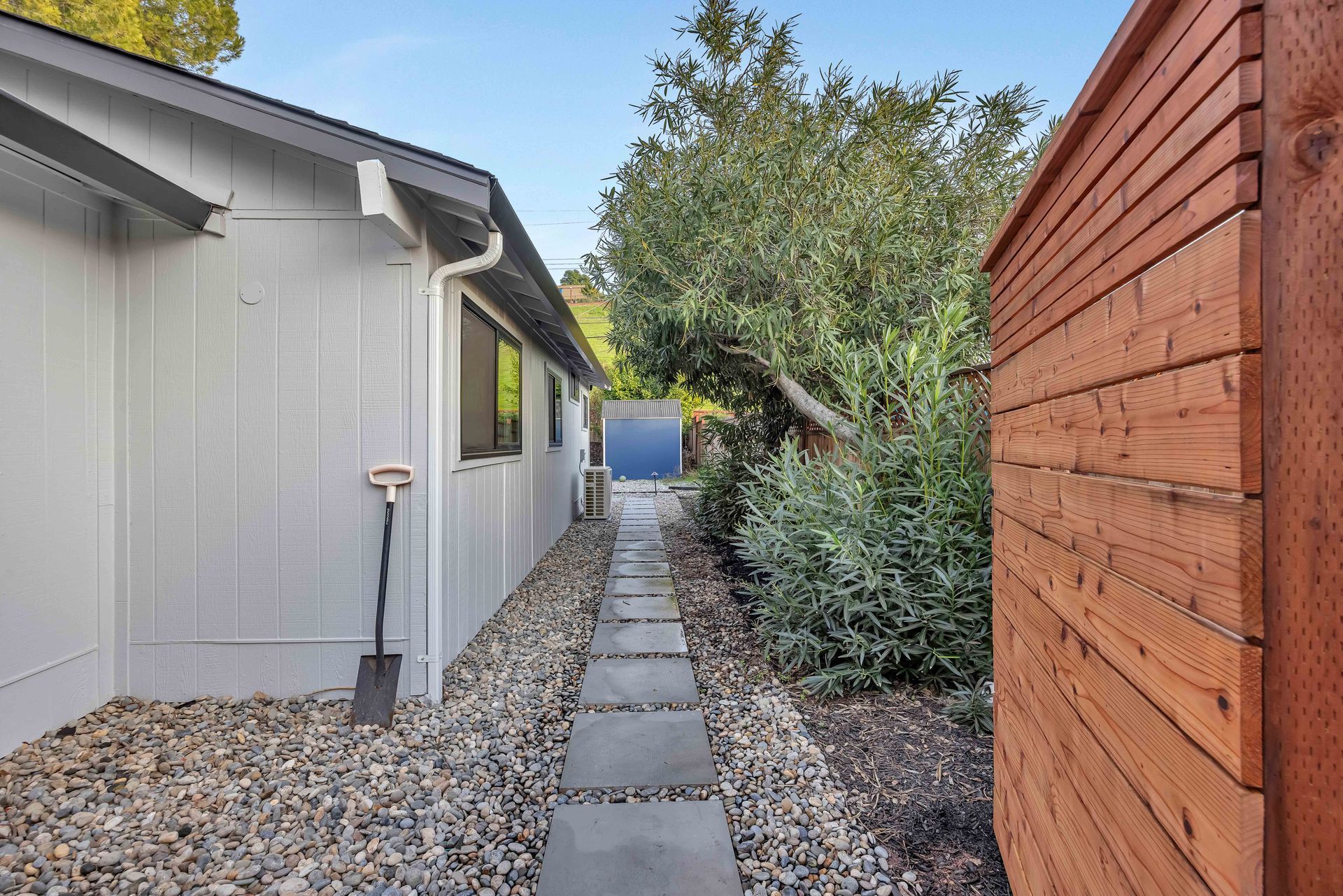 Narrow stone path between a white house and a wooden fence, with gravel and greenery.