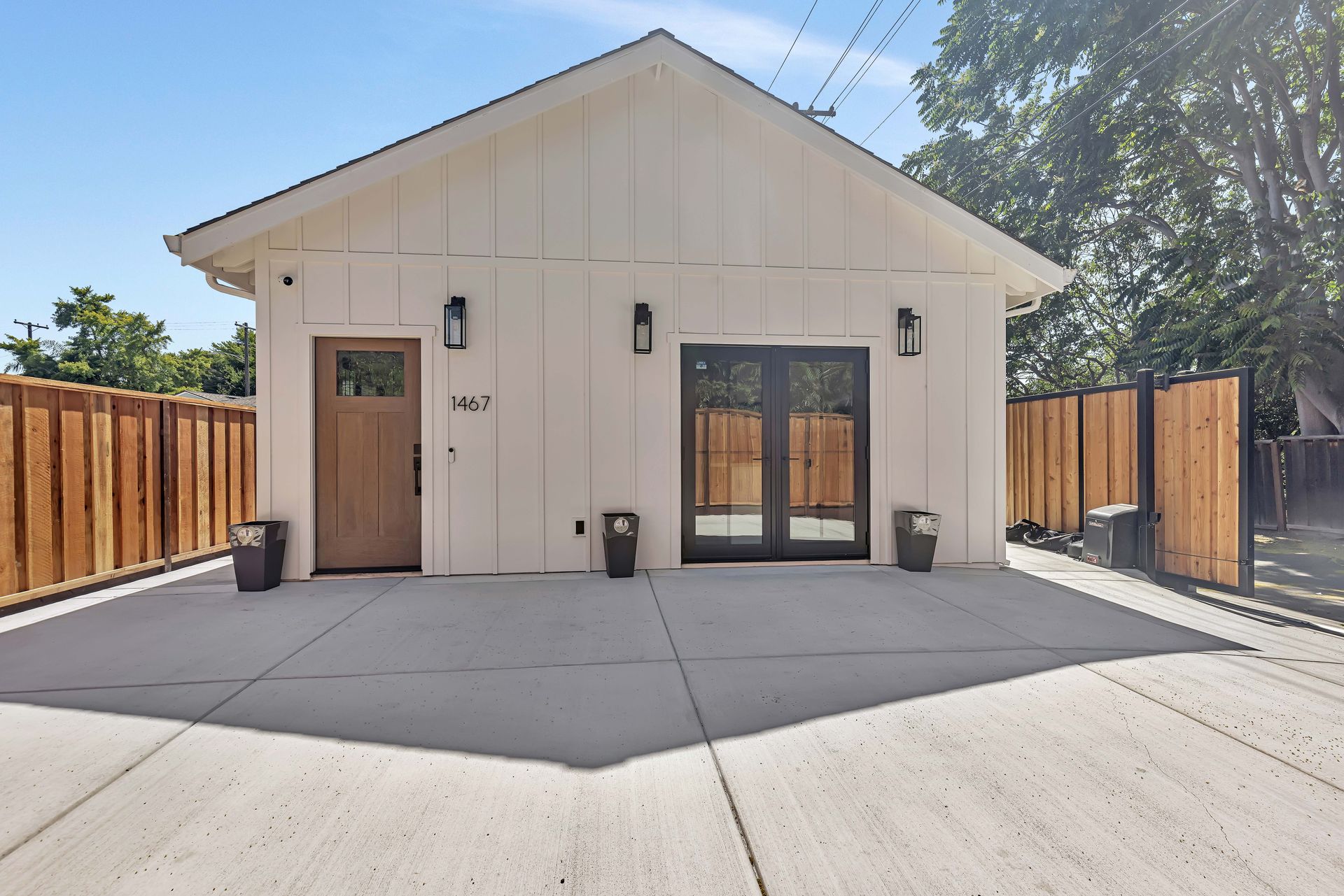 White building with brown door, black double doors, and concrete driveway. Wooden fence surrounds the property.