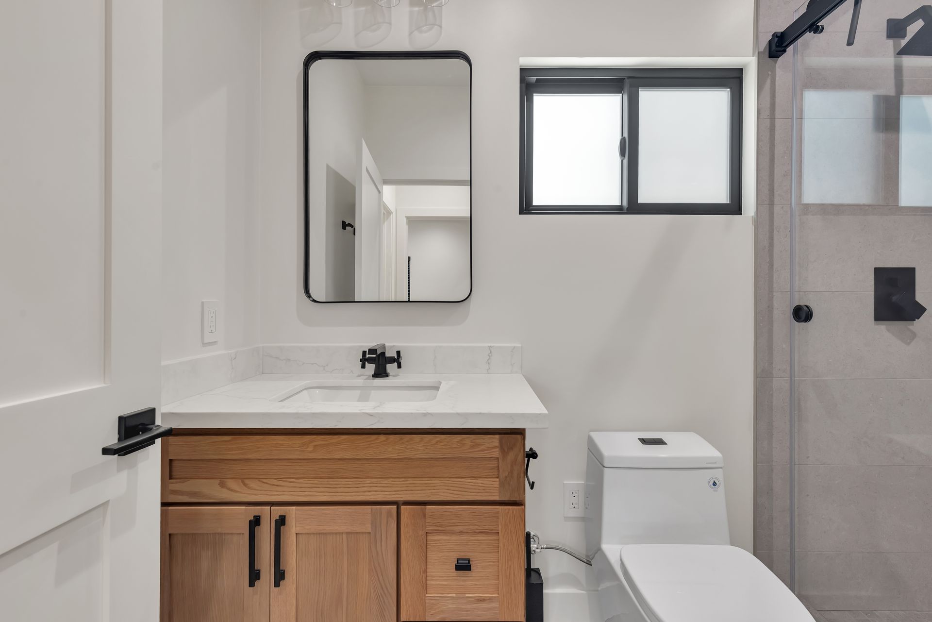 Bathroom with wooden vanity, black-framed mirror, white countertop, toilet, and shower with gray tiles.