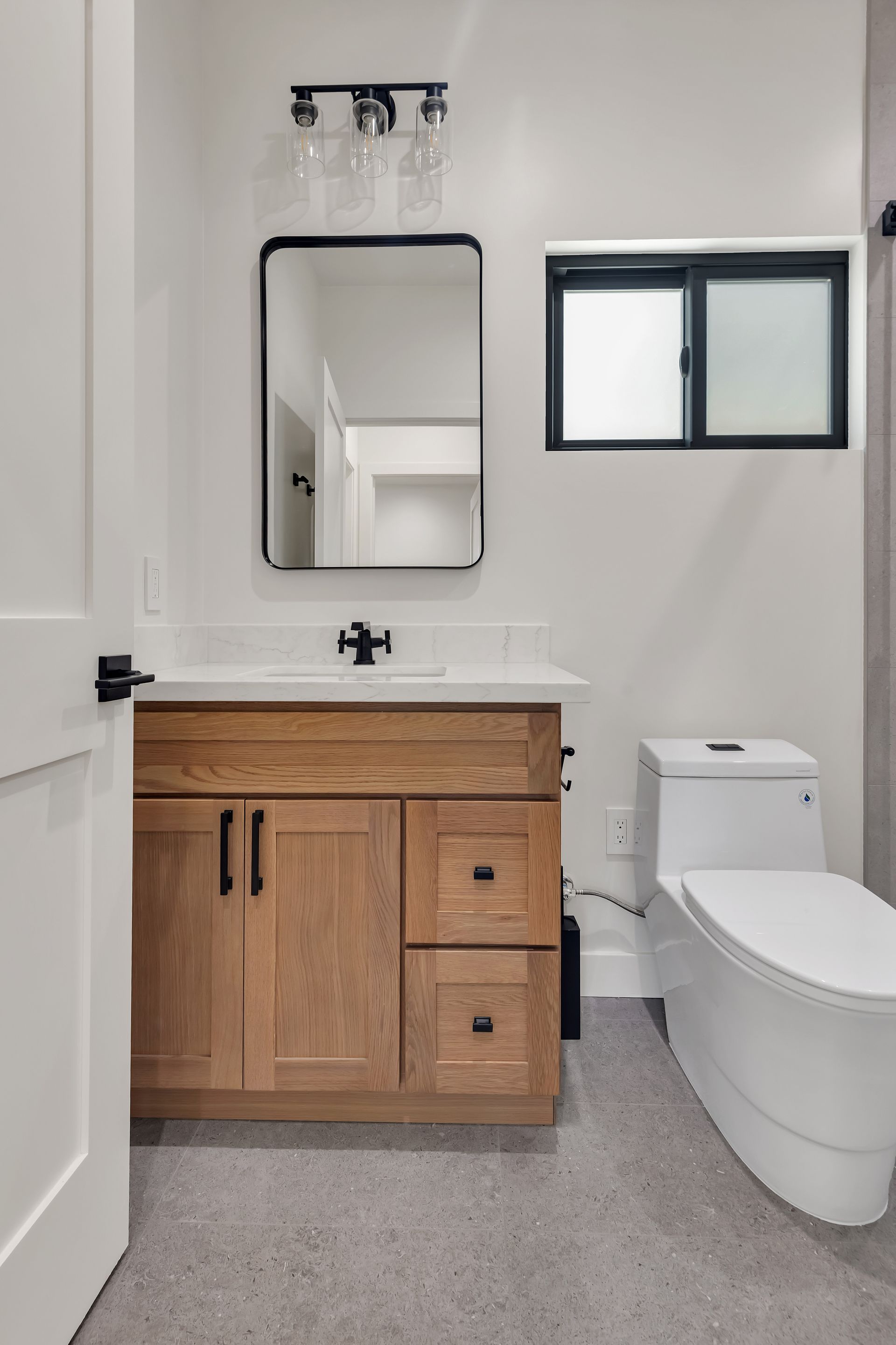 Bathroom with wooden vanity, white toilet, black-framed mirror and window, gray floor, white walls.