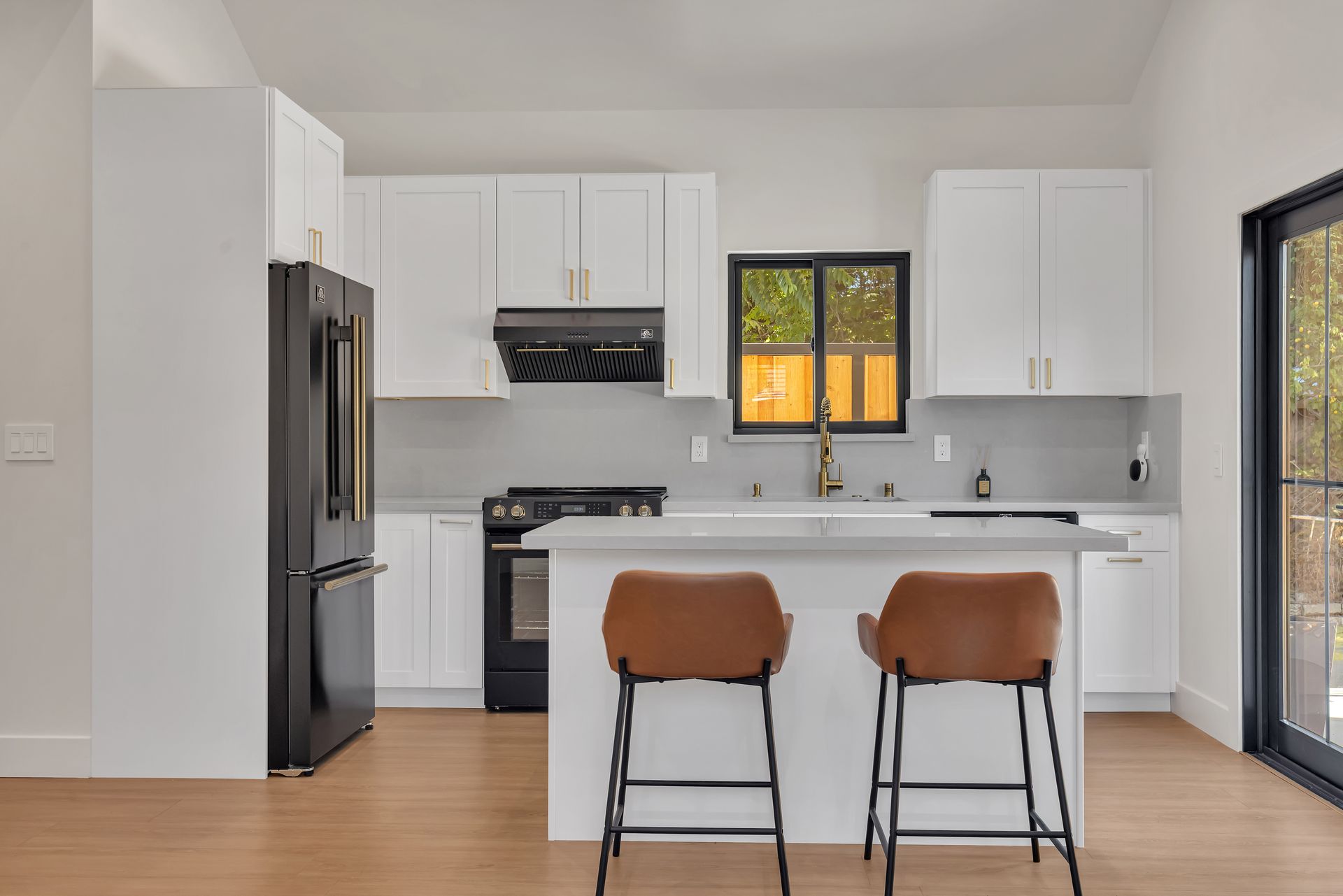 Modern white kitchen with black appliances, a small island with brown bar stools, and a sliding glass door.