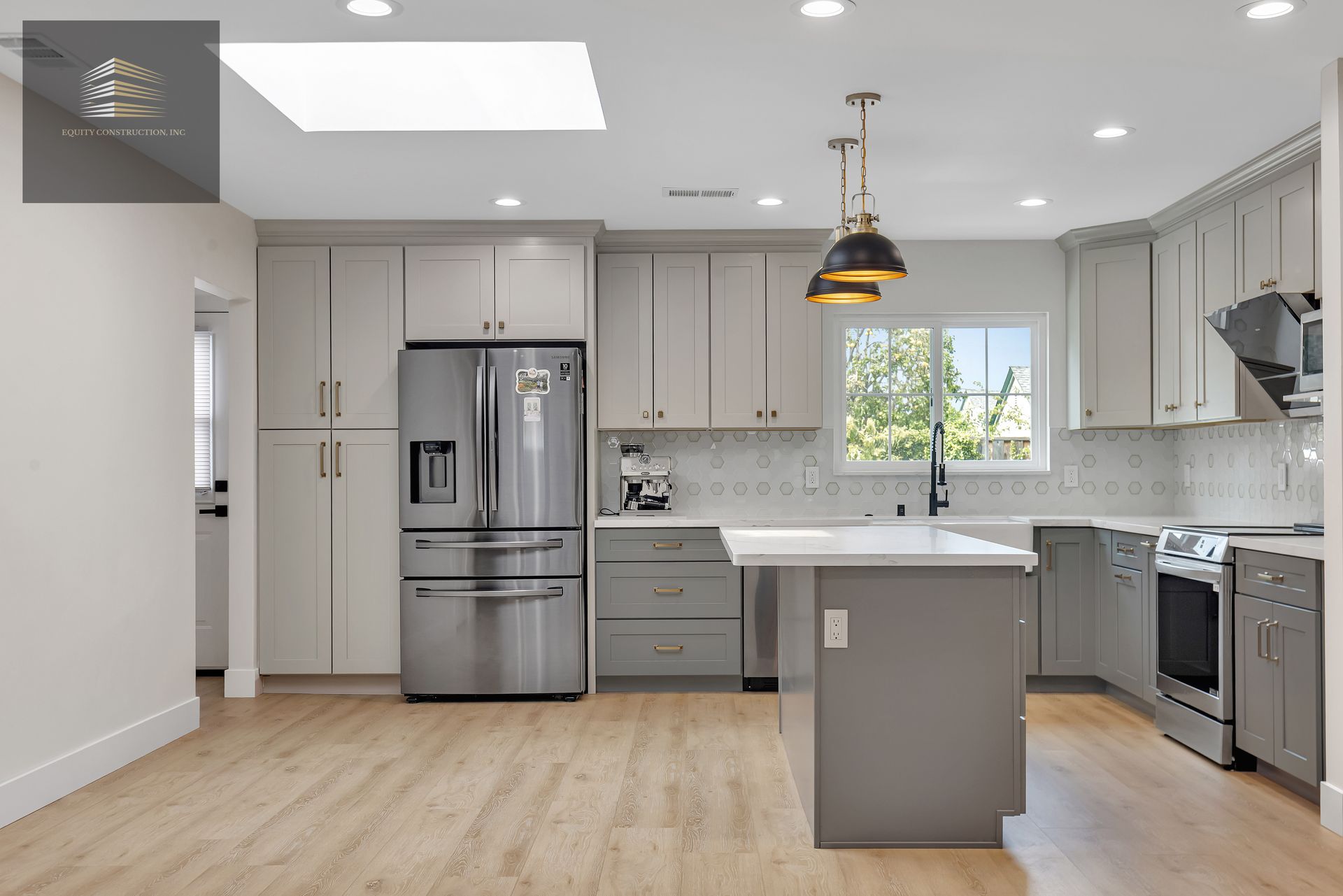 Modern kitchen with gray cabinets, stainless steel appliances, and island. Natural light floods the room.