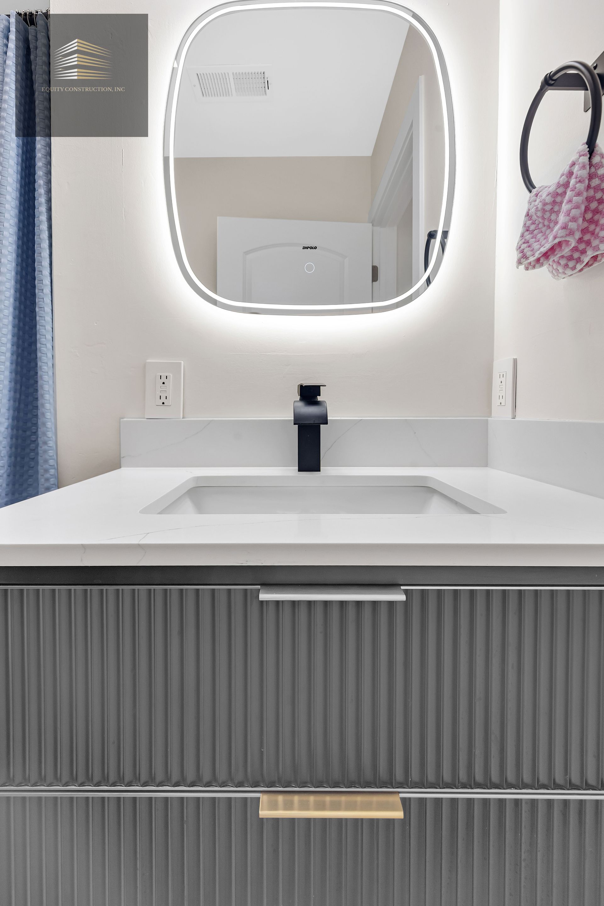 Modern bathroom vanity with a gray ribbed cabinet, white countertop, black faucet, and oval backlit mirror.