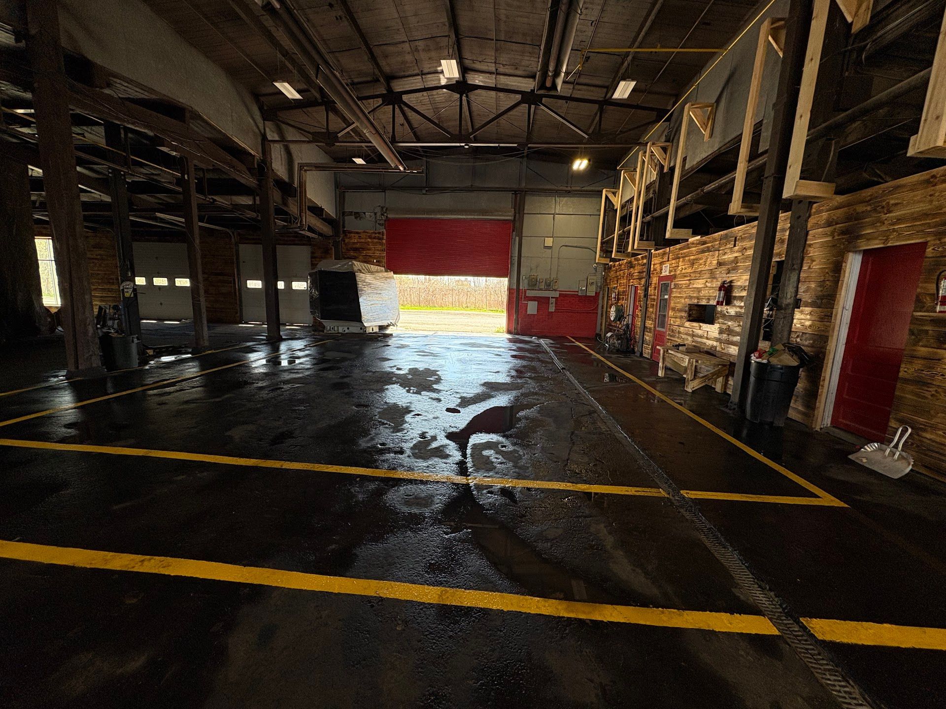 Interior of a wet fire station garage, open door with red door, black floor with yellow lines, wooden beams, bricks, and a hose. 