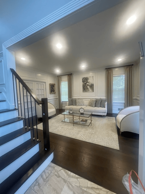 Living room with dark wood floors, a white rug, sofa, and chairs; stairs on the left.