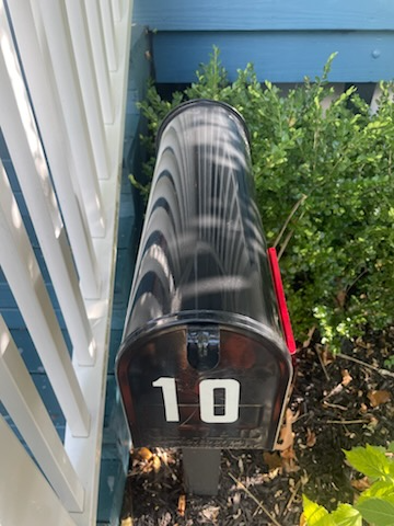 Black mailbox with the number 10 in white, next to a white fence and green bush. Red flag is up.