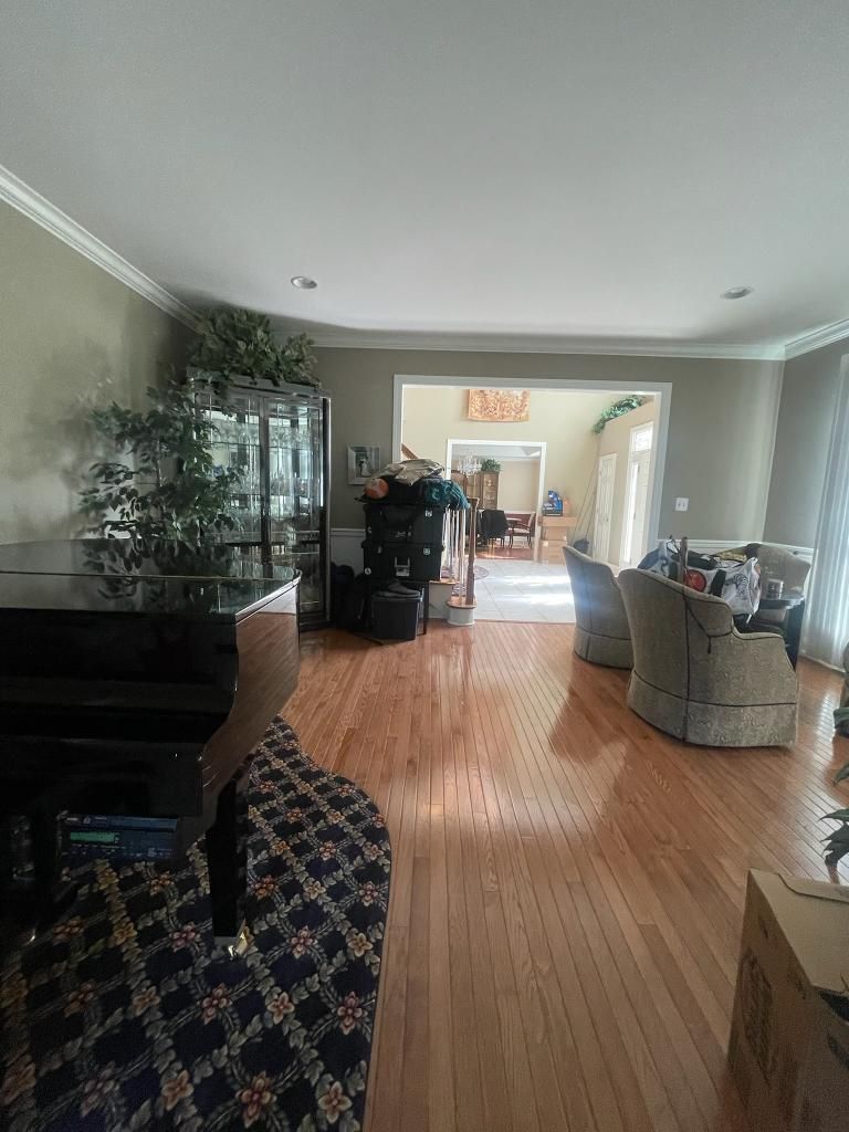 A living room with a piano, plant, cabinet, and chairs, featuring hardwood floors and beige walls.
