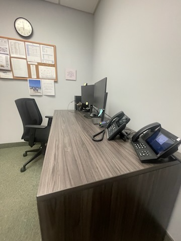 Office desk with two monitors, two phones, black chair, and a bulletin board.