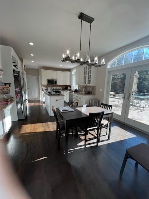 Kitchen with dark wood floors, white cabinets, and a dining table. Sunlight streams in from a glass door.