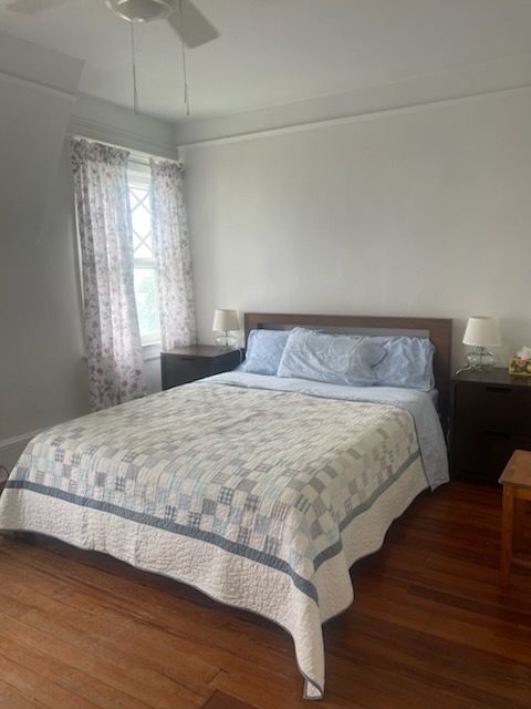 Bedroom with a bed, nightstands, window, and wooden floor. Light-colored bedding and floral curtains.