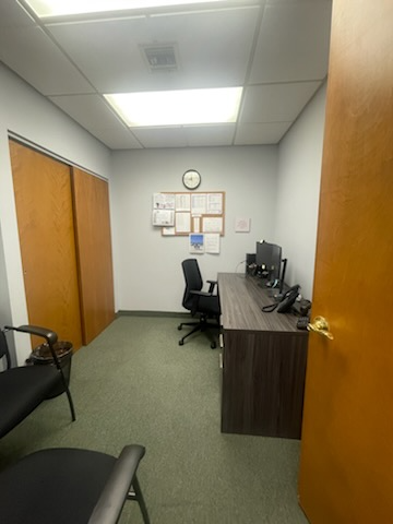 Small office with desk, computer monitors, chair, bulletin board, and two doors. Green carpet, white walls, and brown desk.