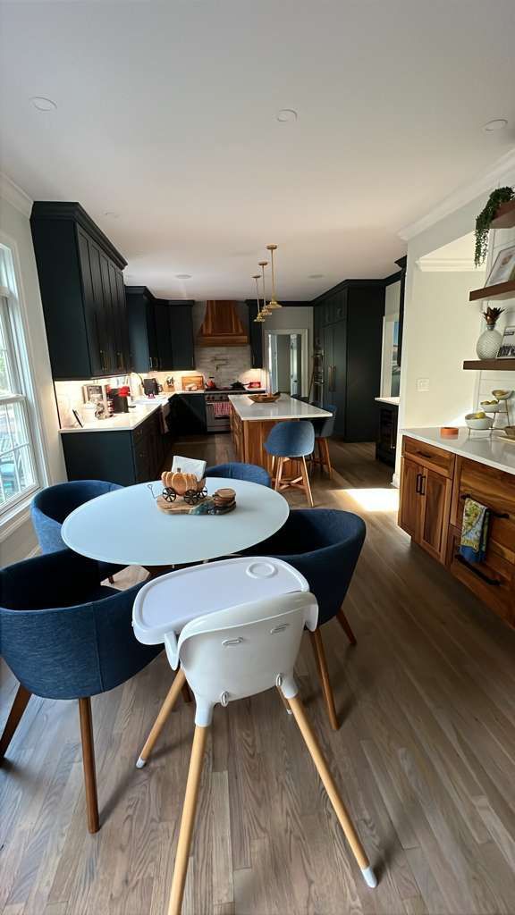 Modern kitchen with dark cabinets, round table, blue chairs, and a high chair; light wood floors.