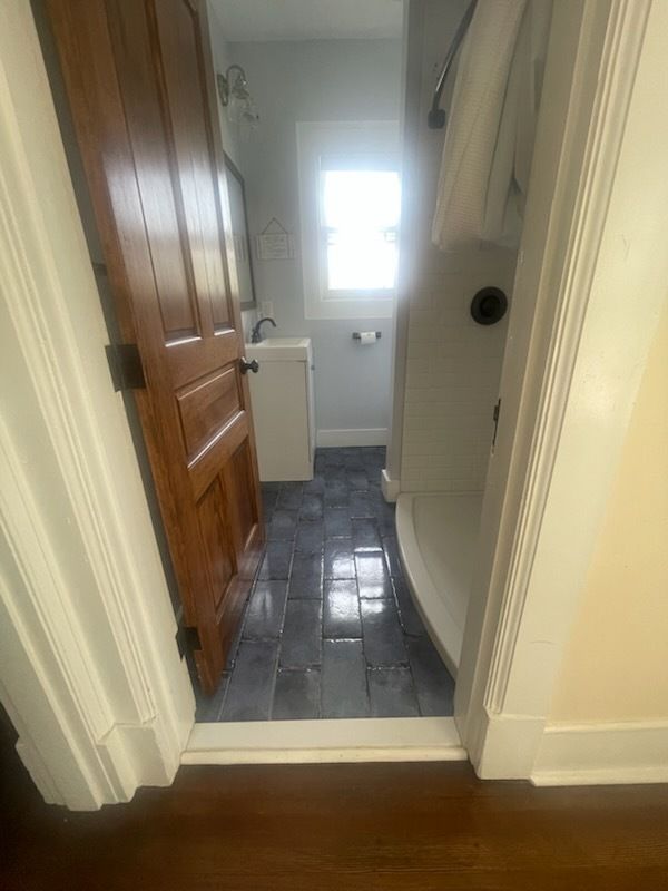Bathroom doorway view. Dark blue tiled floor, wooden door, white fixtures, small window.