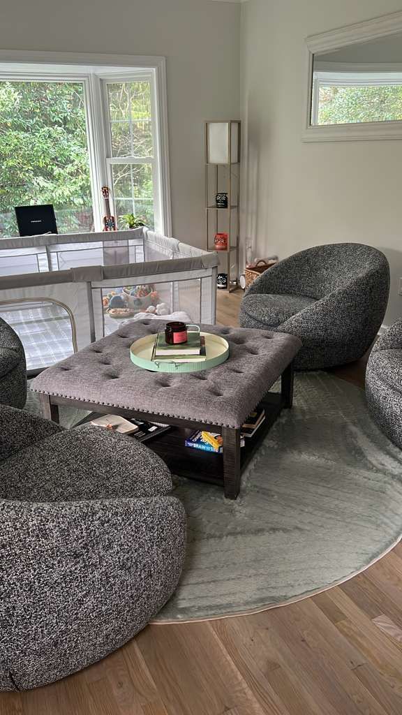 Living room with tufted ottoman, gray chairs, round rug, and a window with greenery.