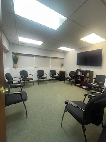 A room with black chairs arranged in a circle, likely a meeting or therapy space. Green carpet and a TV on the wall.