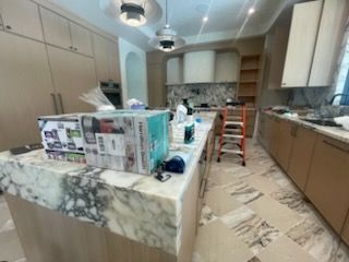 Kitchen renovation in progress: marble island, light wood cabinets, step ladder, appliances, boxes, and various tools.