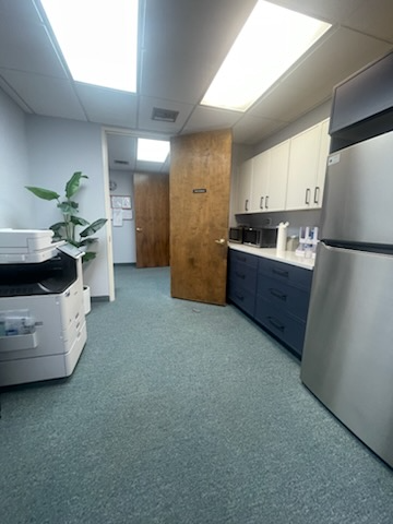 Office interior: copy machine, kitchenette with cabinets, open doors, and blue carpet.