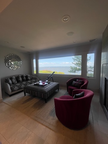 Living room with water view; gray sofa, red chairs, striped ottoman, round mirror, and large window.