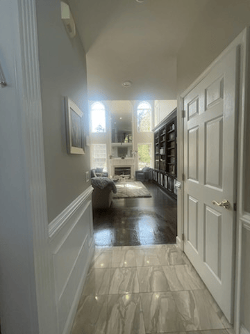 Hallway view into a bright living room with fireplace, bookshelves, and tall windows; white and gray tones.