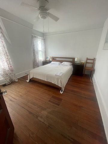 Bedroom with wood floors, queen bed, nightstands, chair, window with floral curtains, and ceiling fan.