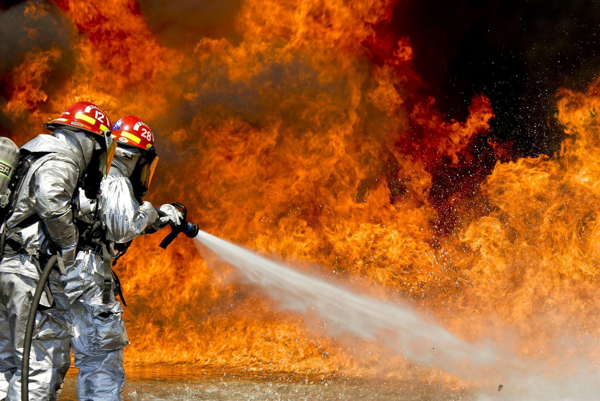 Firefighters in protective gear spraying water at a large, raging fire.