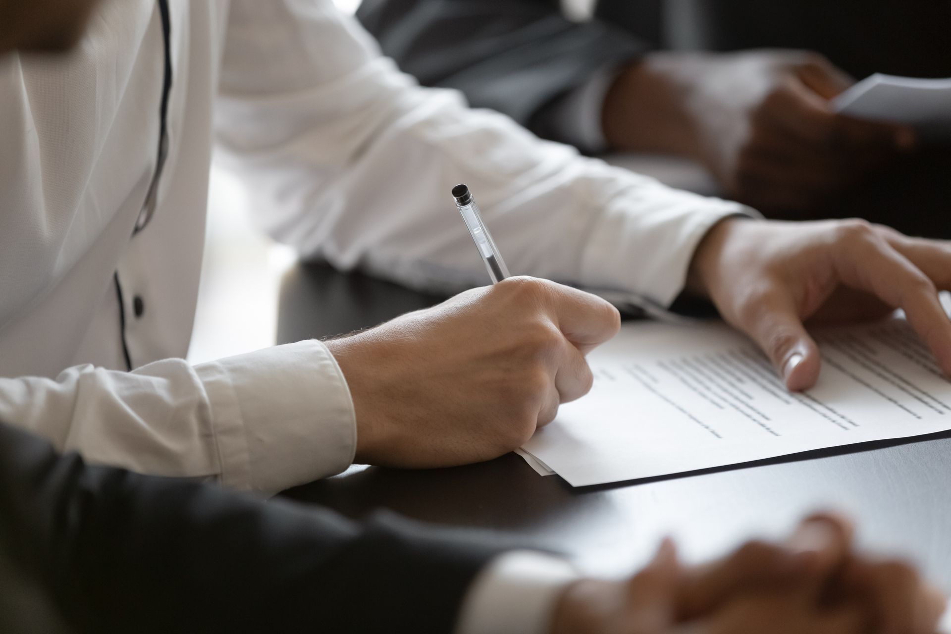 Person signing a document with a pen at a desk, surrounded by others, formal setting.