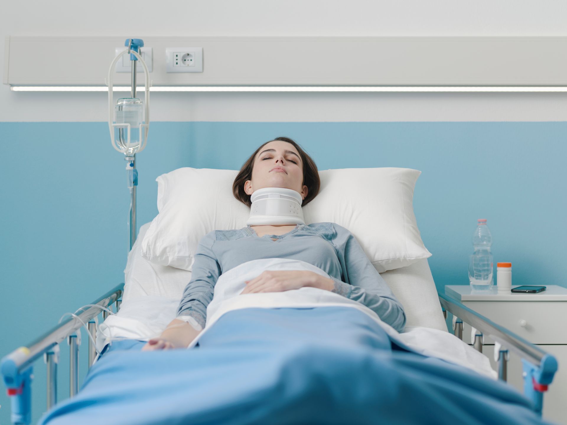 Woman lying in hospital bed, wearing a neck brace, with IV drip and medication on bedside table.