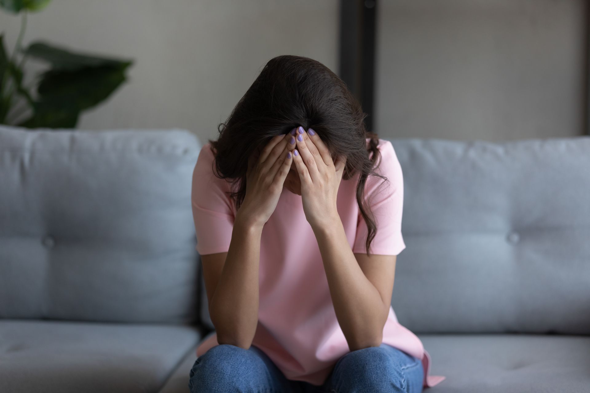 Woman sitting on a couch with hands covering her face, appearing distressed. Pink shirt, blue jeans.