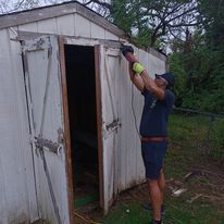 A man is standing in front of a white shed holding a drill.