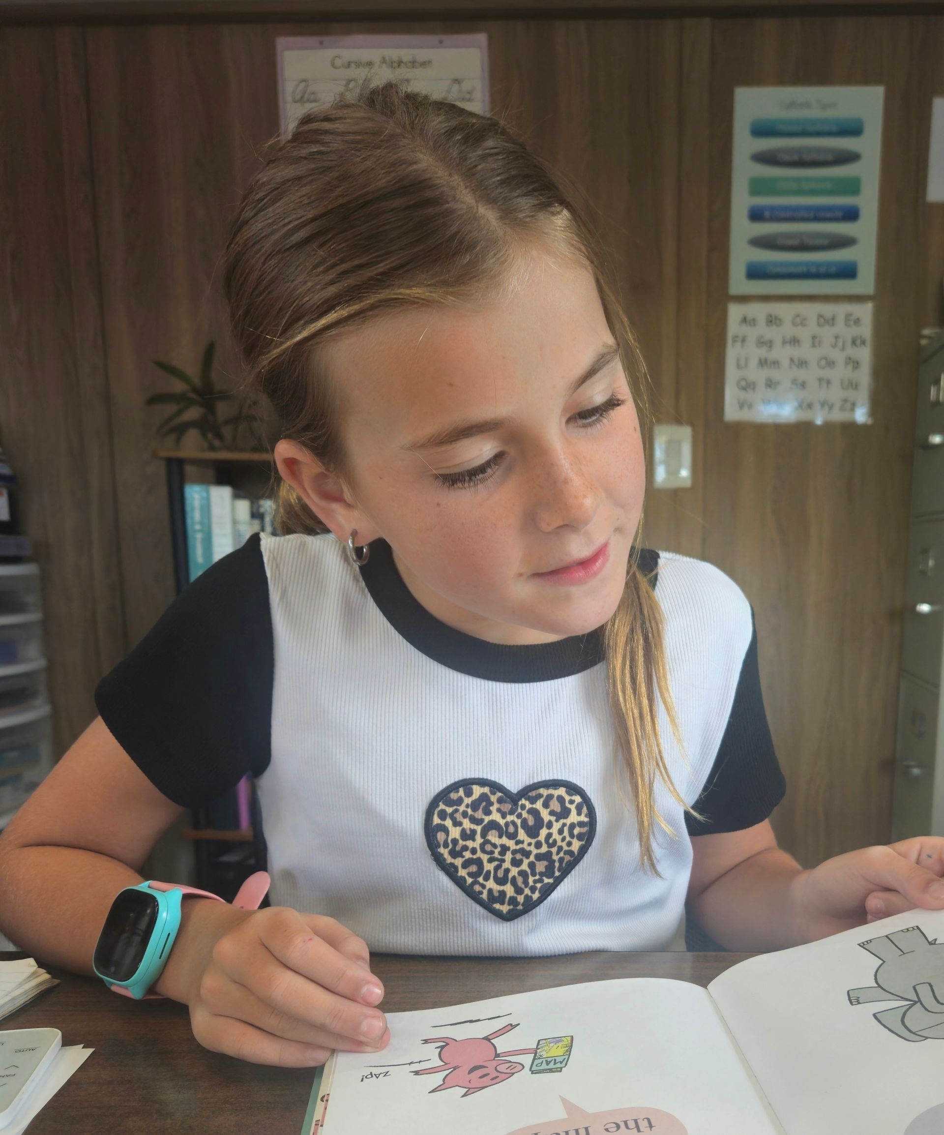 Young girl reading a book indoors, wearing a white shirt with a leopard heart.
