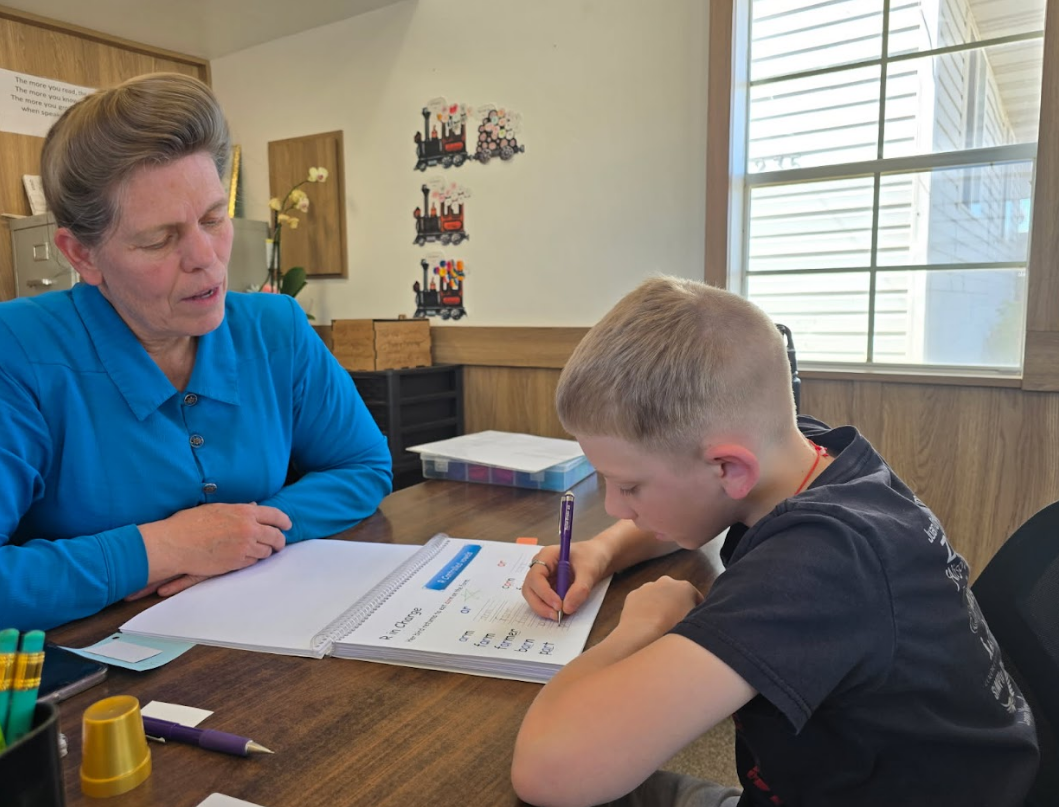 Woman tutoring a young boy at a desk. They are both looking at a notebook. The boy is writing.