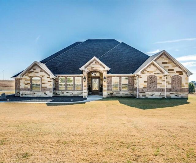 A large brick house with a black roof is sitting in the middle of a grassy field.
