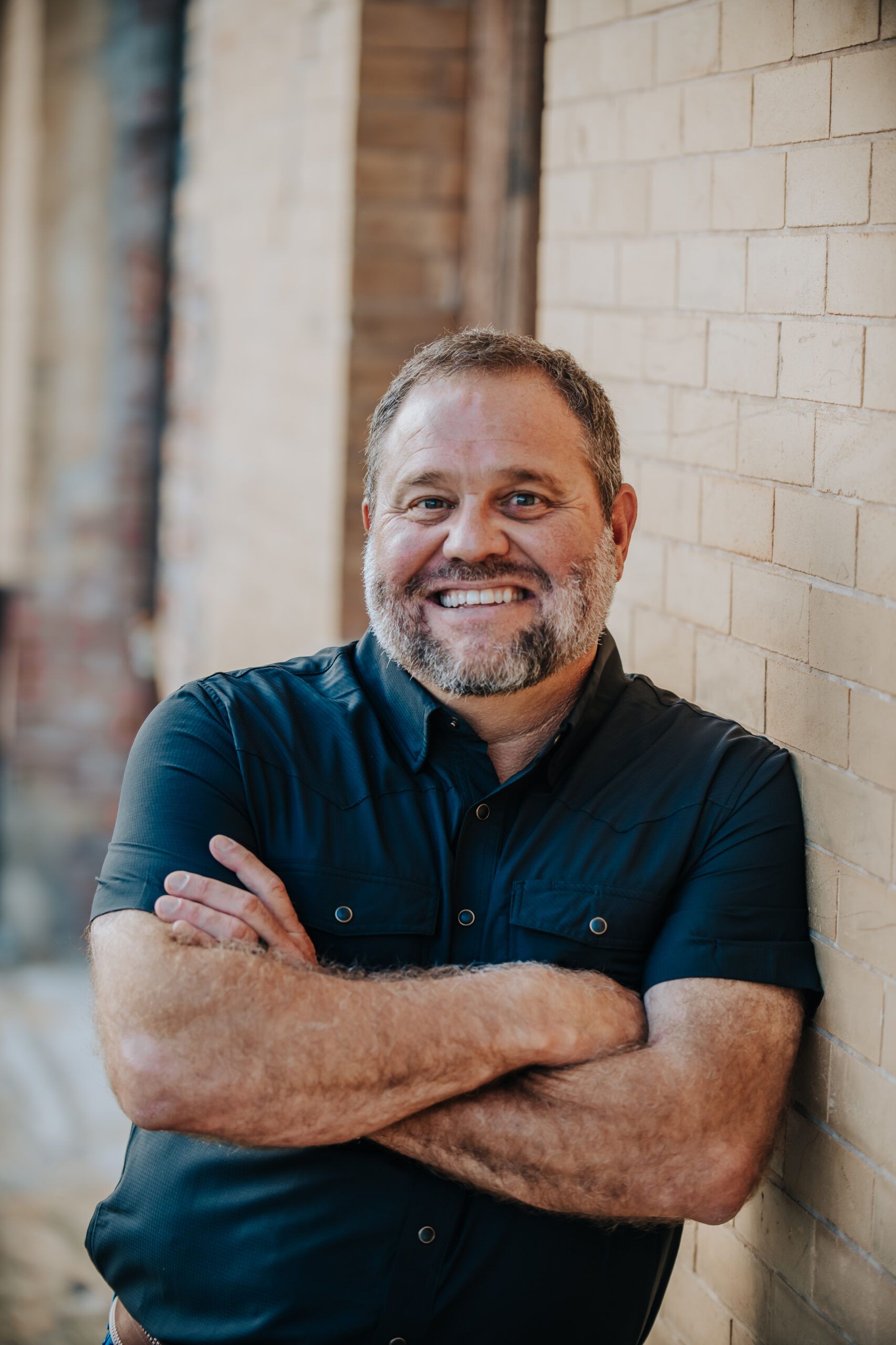 A man with a beard is leaning against a brick wall with his arms crossed and smiling.