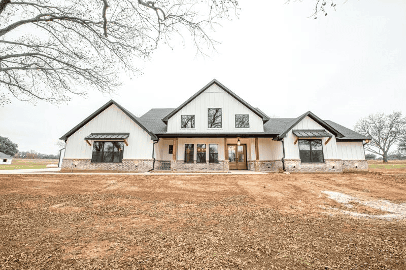 A large white house with a black roof is sitting on top of a dirt field.