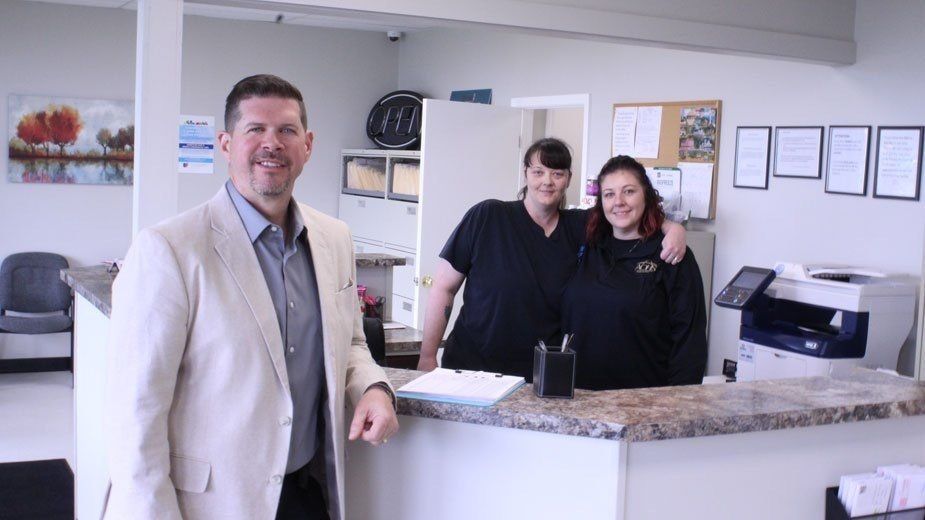 Man in blazer smiles at camera, two women stand behind office counter.