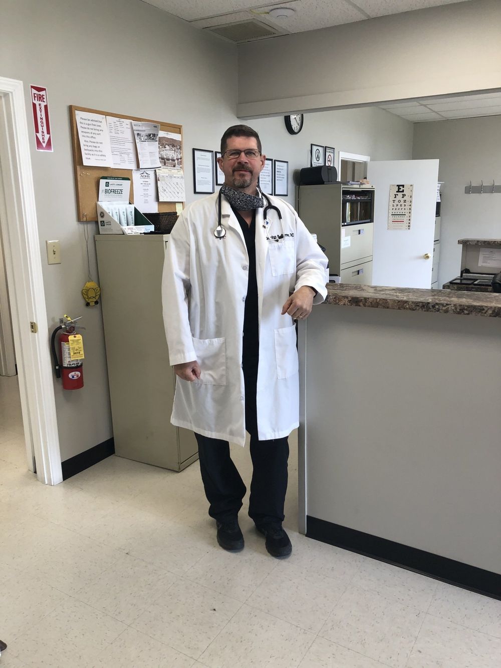 Doctor wearing a white coat and stethoscope, leaning on a counter in an office.