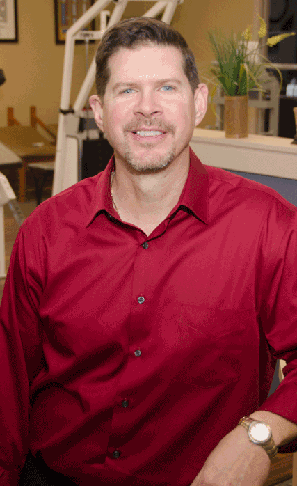 Man in a red button-down shirt smiles, posing indoors near exercise equipment; warm lighting.