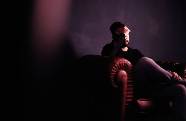 Man sits on a dark red sofa, hand on face, in a dimly lit room with purple hues.