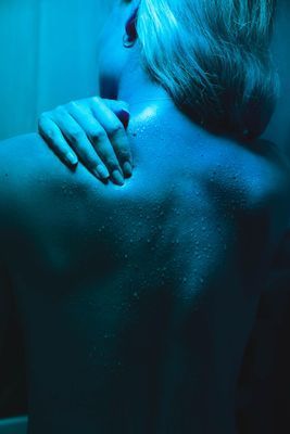 Woman's back, wet with water, hand touching her shoulder in a blue-lit shower.