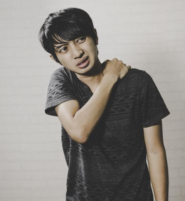 Man holding his shoulder, looking pained. Wearing a dark gray shirt against a white backdrop.