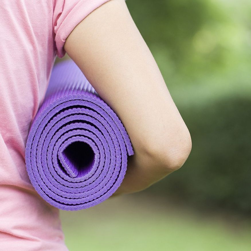 Person in pink shirt carrying a rolled-up purple yoga mat outside.