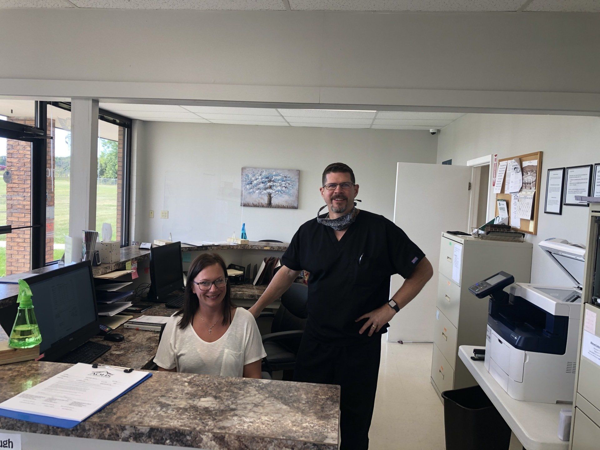 Reception area with two people: woman seated, man standing. Both smiling, behind a counter with a computer and printer.