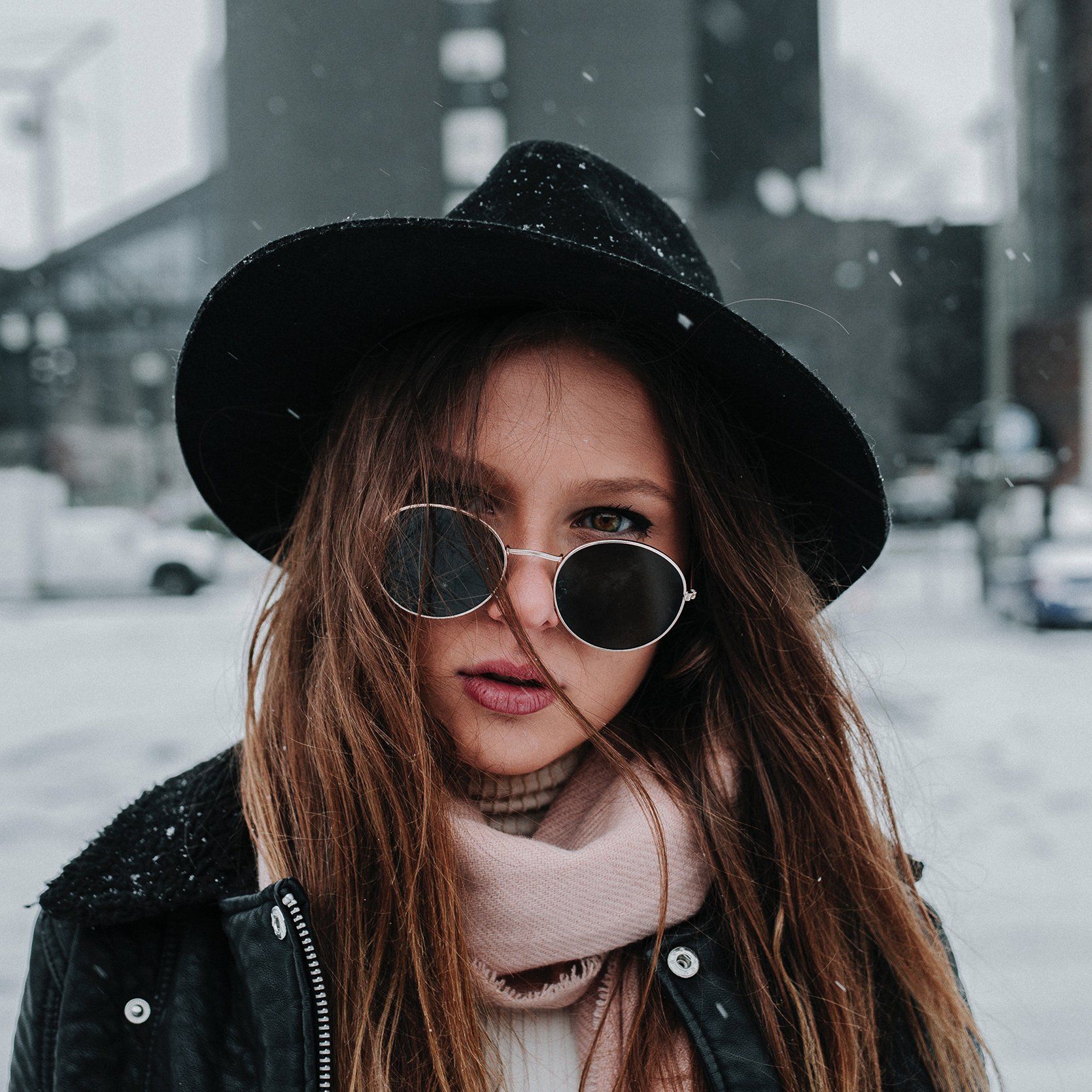 Woman wearing sunglasses, black hat and jacket, in a snowy urban setting.